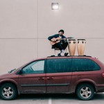Aladean Kheroufi seated on top of a red Dodge Caravan, playing guitar with two conga drums beside him.