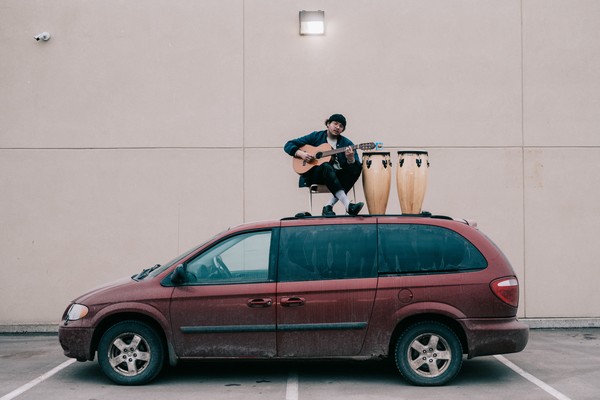 Aladean Kheroufi seated on top of a red Dodge Caravan, playing guitar with two conga drums beside him.