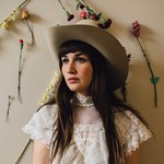 A colour phot shows artist Amy Nelson standing against a beige wall wearing a white dress and a cowboy hat. Flowers are pinned on the wall behind them. 