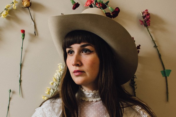 A colour phot shows artist Amy Nelson standing against a beige wall wearing a white dress and a cowboy hat. Flowers are pinned on the wall behind them. 