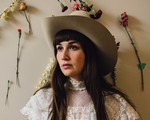 A colour phot shows artist Amy Nelson standing against a beige wall wearing a white dress and a cowboy hat. Flowers are pinned on the wall behind them. 