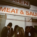 Photo of trio Autolysis standing underneath a sign that reads "MEAT & SAUSAGE" as each member looks toward the ground.