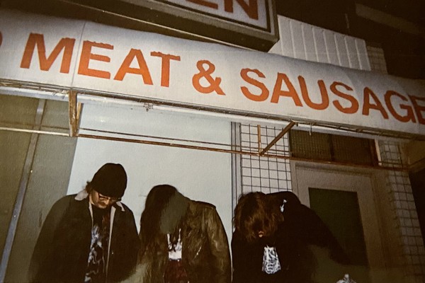 Photo of trio Autolysis standing underneath a sign that reads "MEAT & SAUSAGE" as each member looks toward the ground.