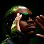 Close up portrait of rapper Banggz in dark sunglasses, looking to the side with his hands on each side of his face.