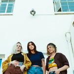 A colour photo taken from below shows three-piece band Barbara from the waist up, standing outside in front of a white building with blue windows and a blue door.  