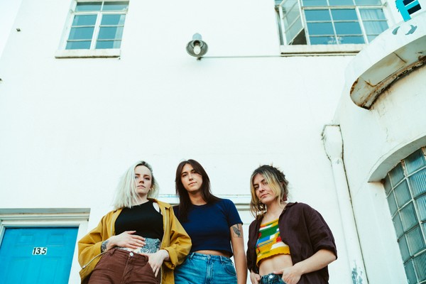 A colour photo taken from below shows three-piece band Barbara from the waist up, standing outside in front of a white building with blue windows and a blue door.  