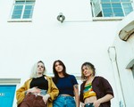 A colour photo taken from below shows three-piece band Barbara from the waist up, standing outside in front of a white building with blue windows and a blue door.  