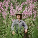 Bennett Mitchell sits in a field of pink flowers and tall grass, looking toward the camera in a brown cowboy hat.