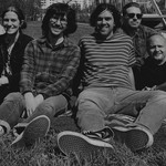 Black and white image of six-piece Big Evil Rat all sitting on a towel in a grassy inner-city park and smiling toward the camera.