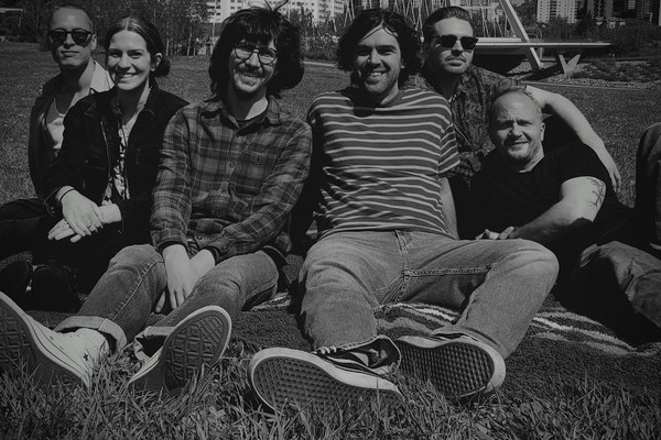 Black and white image of six-piece Big Evil Rat all sitting on a towel in a grassy inner-city park and smiling toward the camera.