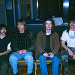 A grainy colour photo shows the four-piece band blind commentary. Each member is seen sitting in a chair, all in a row. 