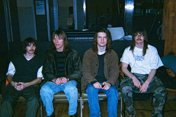 A grainy colour photo shows the four-piece band blind commentary. Each member is seen sitting in a chair, all in a row. 