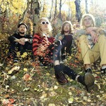 Photo taken in a forest setting in autumn, of fourpiece Cheer. Each member is sitting in the grass.