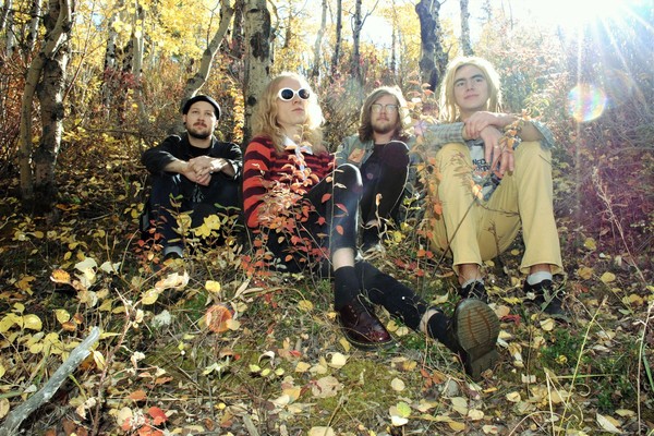 Photo taken in a forest setting in autumn, of fourpiece Cheer. Each member is sitting in the grass.