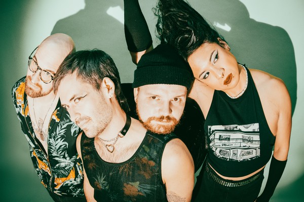 A colour photo of the four members of Chinese Medicine posed closely together against a green background with dramatic lighting casting strong shadows. One member with long black hair lifts it above their head. One member is wearing a black toque. Another wears a mesh tank top and choker necklace, while another member in glasses and a tropical shirt looks upward.
