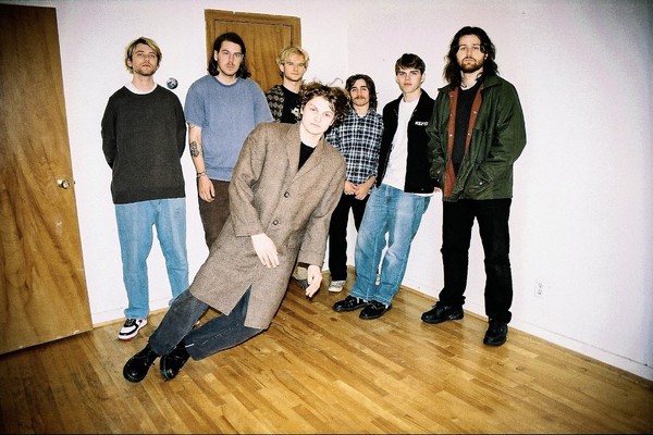 Indoor photo of seven members of Computer are looking toward the camera with one member balancing unusally sideways.