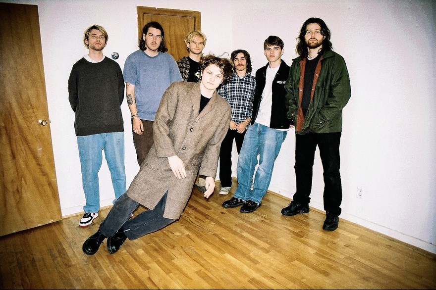 Indoor photo of seven members of Computer are looking toward the camera with one member balancing unusally sideways.
