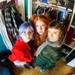A colour photo taken from above shows the three-piece Crachat looking up at the camera, standing in a walk-in closet with clothes hanging behind them. 