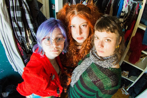 A colour photo taken from above shows the three-piece Crachat looking up at the camera, standing in a walk-in closet with clothes hanging behind them. 