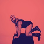 A colour photo shows artist Devours on a black stool on their hands and knees in a pink room. They are wearing a one-piece outfit and cowboy boots. 