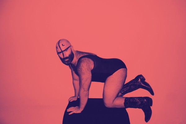 A colour photo shows artist Devours on a black stool on their hands and knees in a pink room. They are wearing a one-piece outfit and cowboy boots. 