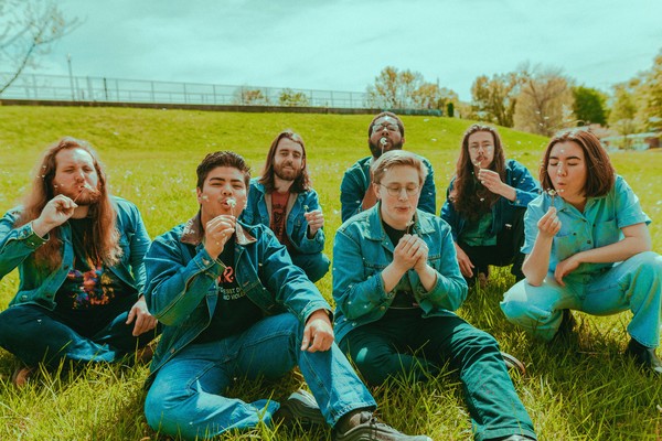 Outdoor photo taken on a field on a sunny day, featuring seven members of DOOM GONG blowing dandelions while dressed in matching denim