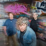 A colour photo of the three members of Down The Lees standing in front of a graffiti-covered wall with motion blur effects. The central figure, with curly hair and a denim jacket, leans forward toward the camera with a serious expression. Two other members stand behind, one in a dark button-up shirt and the other in a navy shirt and khakis.