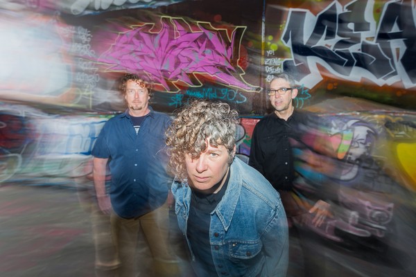 A colour photo of the three members of Down The Lees standing in front of a graffiti-covered wall with motion blur effects. The central figure, with curly hair and a denim jacket, leans forward toward the camera with a serious expression. Two other members stand behind, one in a dark button-up shirt and the other in a navy shirt and khakis.