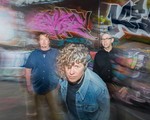 A colour photo of the three members of Down The Lees standing in front of a graffiti-covered wall with motion blur effects. The central figure, with curly hair and a denim jacket, leans forward toward the camera with a serious expression. Two other members stand behind, one in a dark button-up shirt and the other in a navy shirt and khakis.