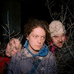 Taken through barren bushes, a photo featuring three members of Dresser, looking off to the side