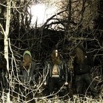 Dark image featuring four members of Feeding, each hooded with long hair and gazing toward the ground as they stand in a barren forest