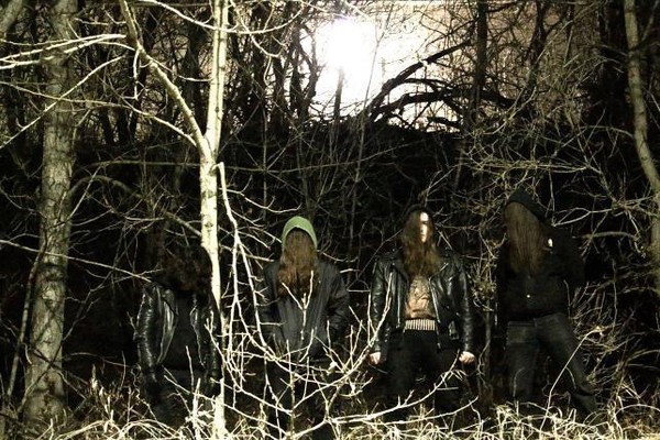 Dark image featuring four members of Feeding, each hooded with long hair and gazing toward the ground as they stand in a barren forest