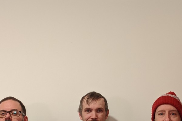 A colour photo shows three-piece band Fulfilment standing in a line against a beige wall. Each member is only shown from the nose up at the bottom of the frame.
