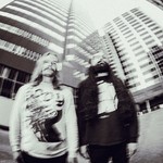 A blurry black and white photo shows two members of Genizah standing outside with two tall buildings behind them.