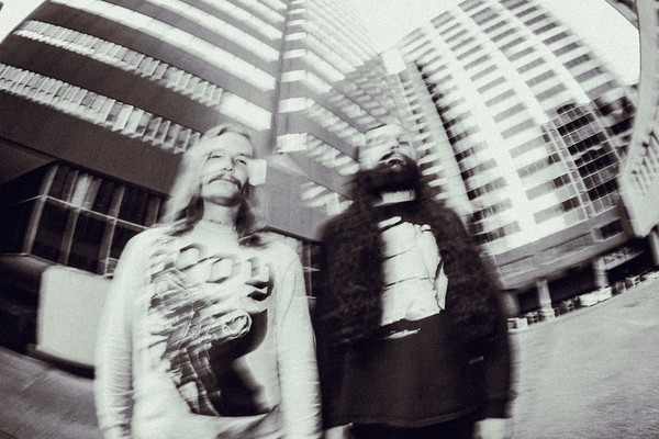 A blurry black and white photo shows two members of Genizah standing outside with two tall buildings behind them.