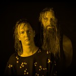 A colour photo shows the duo Grimelda standing against a black background. An effect on the photo makes both members appear yellow.