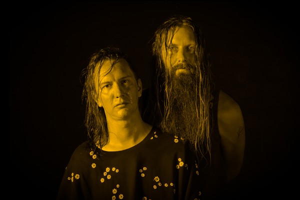 A colour photo shows the duo Grimelda standing against a black background. An effect on the photo makes both members appear yellow.