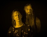 A colour photo shows the duo Grimelda standing against a black background. An effect on the photo makes both members appear yellow.