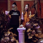 A colour photo of the duo HAIR CONTROL shows them standing in a room with fake plants and a white pillar in the middle with a conch shell sitting on top. The member on the left wears white sunglasses and holds a conch shell. Their black t-shirt says CULT MEMBER. The member on the right is in an orange outfit and hold a mallet in their hand.