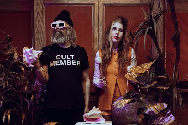 A colour photo of the duo HAIR CONTROL shows them standing in a room with fake plants and a white pillar in the middle with a conch shell sitting on top. The member on the left wears white sunglasses and holds a conch shell. Their black t-shirt says CULT MEMBER. The member on the right is in an orange outfit and hold a mallet in their hand.
