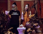 A colour photo of the duo HAIR CONTROL shows them standing in a room with fake plants and a white pillar in the middle with a conch shell sitting on top. The member on the left wears white sunglasses and holds a conch shell. Their black t-shirt says CULT MEMBER. The member on the right is in an orange outfit and hold a mallet in their hand.