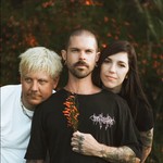 A colour photo shows three-piece band HATERS standing close together outside in front of a bush with orange flowers. The center person is holding an orange flower from the bush in front of their chest. 