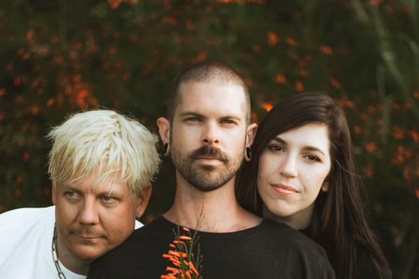 A colour photo shows three-piece band HATERS standing close together outside in front of a bush with orange flowers. The center person is holding an orange flower from the bush in front of their chest. 