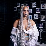 A colour photo of artist HAWA B standing inside a black tile bathroom with polaroids taped on the wall behind them.