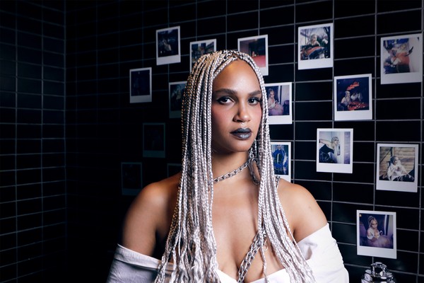 A colour photo of artist HAWA B standing inside a black tile bathroom with polaroids taped on the wall behind them.