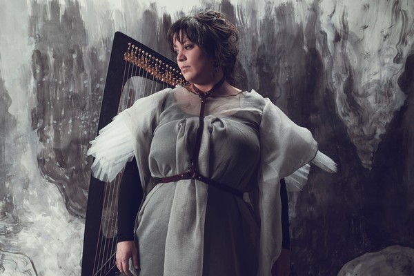 A colour photo shows artist Hermitess standing in a grey painted room in a grey dress with a harp sitting behind them. 