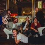 A colour photo shows six-piece band Hip Hip Thursday posing outside of a venue. One member on the left is holding a bass, and another in the middle is holding both a trumpet and a saxaphone.