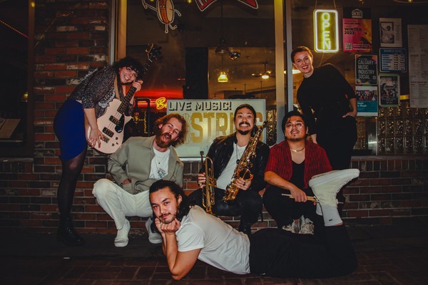 A colour photo shows six-piece band Hip Hip Thursday posing outside of a venue. One member on the left is holding a bass, and another in the middle is holding both a trumpet and a saxaphone.