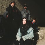 A colour photo shows five-piece band Holy Void standing outside in front of a grey brick wall. Three member crouch and two stand behind them. There is snow on the ground and all members are bundled up in warm clothes. 