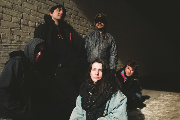 A colour photo shows five-piece band Holy Void standing outside in front of a grey brick wall. Three member crouch and two stand behind them. There is snow on the ground and all members are bundled up in warm clothes. 